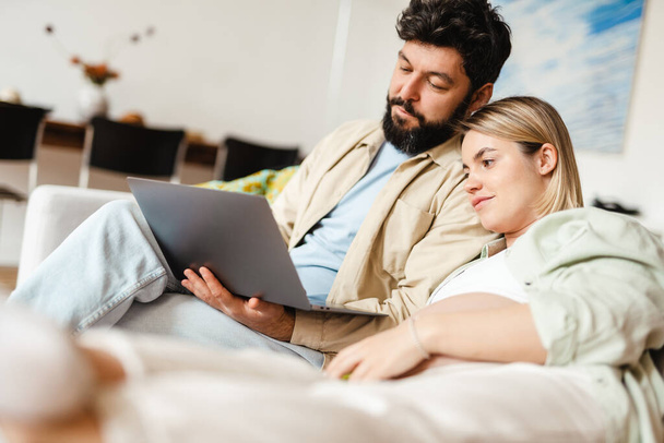 Bearded man and pregnant woman using laptop while sitting on couch at home - Foto, imagen