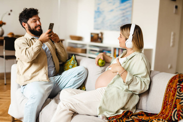 White man taking photo of his pregnant wife while sitting on couch at home - Foto, afbeelding