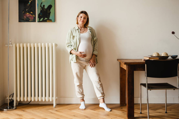 Valkoinen nuori raskaana oleva nainen hymyilee ja katsoo kameraa kotona - Valokuva, kuva