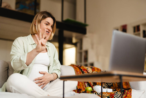 Young pregnant woman taking video call on laptop while sitting on couch at home - Fotoğraf, Görsel
