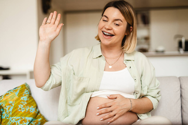 Young pregnant woman laughing and gesturing while sitting on couch at home - Φωτογραφία, εικόνα