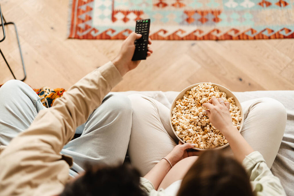 White couple eating popcorn and watching movie while sitting on couch at home - 写真・画像