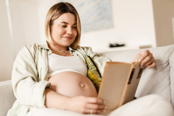 Young pregnant woman smiling and reading book while sitting on couch at home - 写真・画像