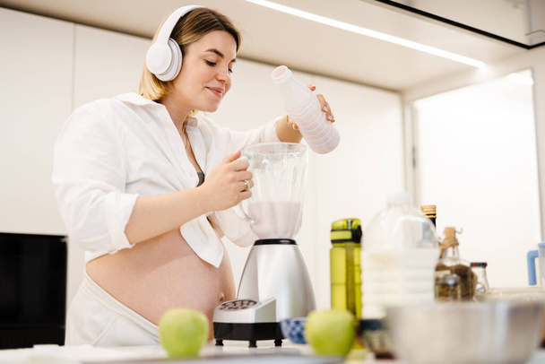 White pregnant woman in headphones making smoothy at home - Fotó, kép