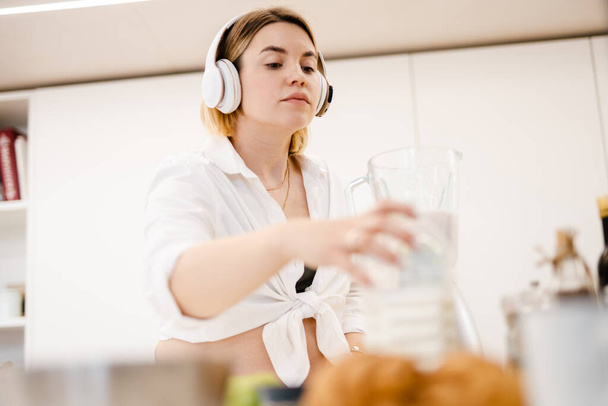 White pregnant woman in headphones making smoothy at home - Photo, Image