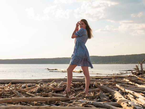 A young beautiful barefoot woman with slender legs in a summer blue sundress dress on the shore of the sea bay at sunset, the concept of a beach holiday, summer walks in nature, an active lifestyle, a weekend at the sea - Foto, Bild