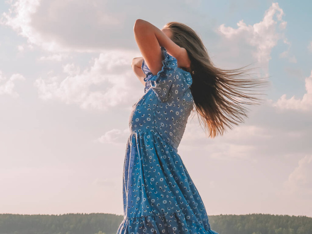 A young blonde woman in a blue dress with her hair fluttering in the wind against the sky on a summer evening, emotions of happiness, freedom and joy, the concept of outdoor recreation, an active lifestyle, traveling and walking in nature,  sunny tan - Foto, immagini