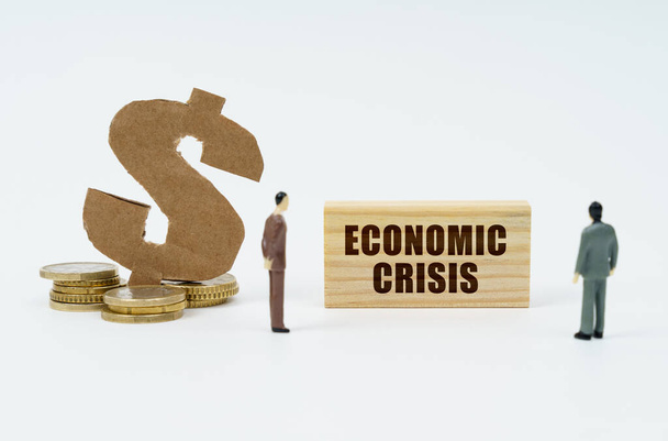 Wirtschafts- und Finanzkonzept. Auf weißem Hintergrund Münzen, ein Dollarsymbol und Figuren von Geschäftsleuten, die eine Holzplatte mit der Aufschrift betrachten - Wirtschaftskrise - Foto, Bild