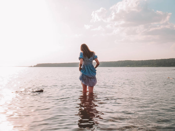 A young beautiful barefoot woman with slender legs in a summer blue sundress dress on the shore of the sea bay at sunset, the concept of a beach holiday, summer walks in nature, an active lifestyle, a weekend at the sea - Valokuva, kuva