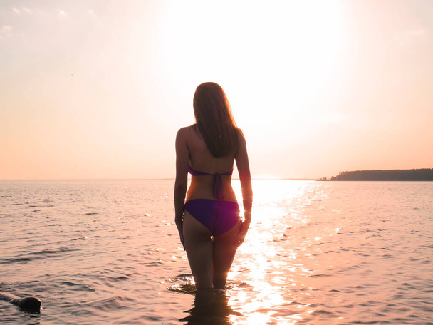 A young woman in a bikini with a slender attractive body on the beach against the background of water in the golden light of the setting sun, the concept of a beach holiday, sun tanning, swimming in the sea, an active and healthy lifestyle - Photo, Image