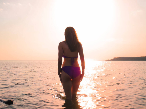 Nuori nainen bikineissä, jossa on hoikka houkutteleva runko rannalla veden taustalla laskevan auringon kultaisessa valossa, rantaloman, aurinkorusketuksen, meressä uimisen, aktiivisen ja terveellisen elämäntavan käsite. - Valokuva, kuva
