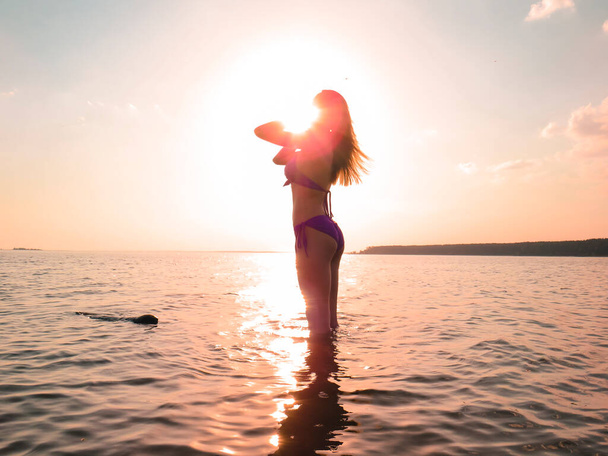 Młoda kobieta w bikini z smukłym, atrakcyjnym ciałem na plaży na tle wody w złotym świetle zachodzącego słońca, koncepcja wakacji na plaży, opalanie się, pływanie w morzu, aktywny i zdrowy tryb życia - Zdjęcie, obraz