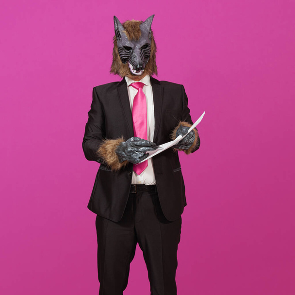 Against a pink background is a man dressed in a black suit with a jacket, white shirt and tie, wearing a werewolf mask, holding a document and pen offering to sign it. - Photo, image