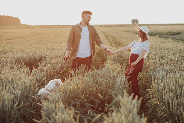 Νεαρή έγκυος γυναίκα και σύζυγος περπατούν στο χωράφι, κρατιούνται χεράκι χεράκι. Ζευγάρι περιμένει το μωρό, χαλαρώστε έξω με το σκυλί - Φωτογραφία, εικόνα