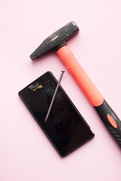 Hammer, nail and mobile phone with broken display on pink background. Crashed smartphone concept. Top view. - Photo, image