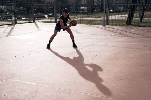 mladý muž hraje basketbal venku na ulici s dlouhými stíny - Fotografie, Obrázek