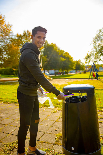 Sonriendo joven voluntario poniendo pajitas en cubo de basura en el parque - Foto, imagen