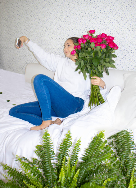 Ein schönes, junges Mädchen mit einem Strauß roter Rosen sitzt auf dem Bett und macht ein Selfie  - Foto, Bild