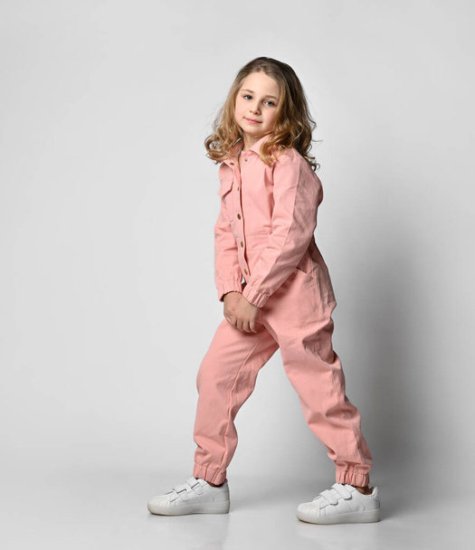 Full length profile picture of a positive curly blonde girl standing in white sneakers and pink cotton overalls, half side on a light background - Photo, image