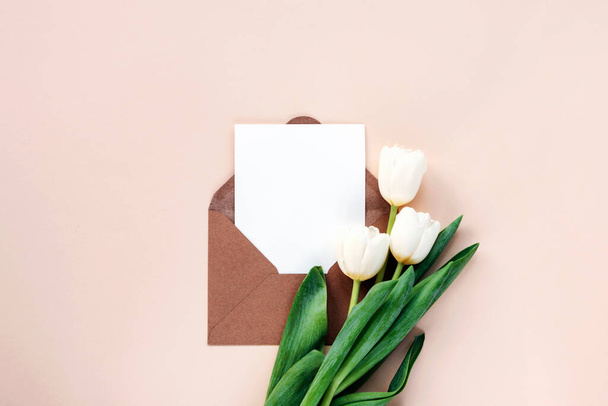 ホワイトチューリップ、ニュートラルベージュの背景にブランクシート付きの封筒。バレンタインデー、母の日の休日の概念。トップビュー、フラットレイアウト、モックアップ. - 写真・画像