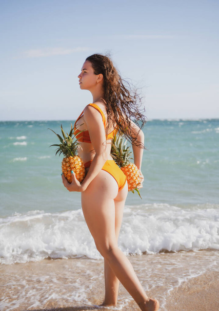 Sexy woman holding fresh pineapple. Buttocks of girl in bikini with fruit pineapple on beach background, summer vacation, healthy food and fitness. - Fotoğraf, Görsel