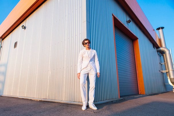 Wide angle portrait of a handsome man dressed in a white suit and round hippie sunglasses standing near the corner of a building during sunset - Foto, immagini