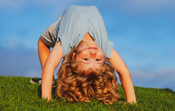 Happy boy doing a headstand on the grass in the summer sunshine. Funny happy little child standing on head upside down on the grass in the park. Childhood concept. - 写真・画像