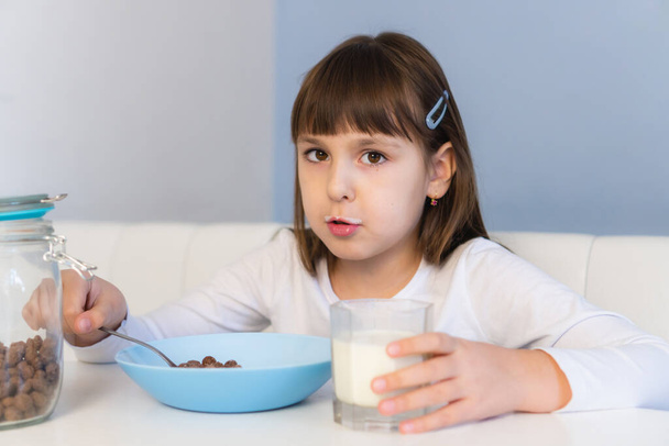 女の子は白いテーブルに座ってミルクとチョコレートコーンボールを食べる。子供は朝食を - 写真・画像