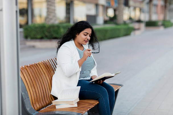 A beautiful girl is sitting on a bench and reading a book. The concept of urban lifestyle and education. Side view. - Foto, Imagen