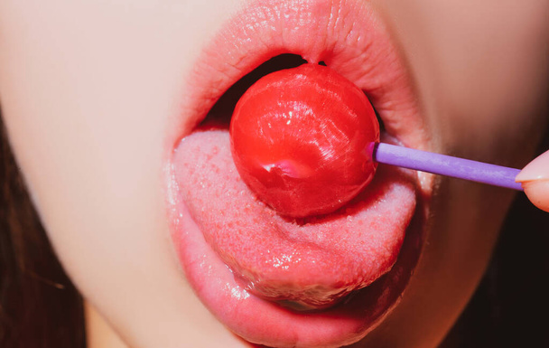 Girl with sexy mouth eating chupa chups close up. Woman lips sucking lollypop. Woman holding lollipop in mouth, close up. Red lips, sensual and sex shop concept. - 写真・画像