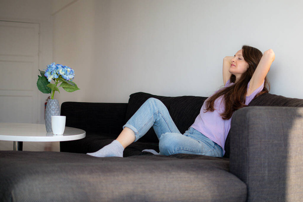 Eine junge Frau sitzt auf einem Sofa in Balkonnähe und dehnt sich am Morgen - Foto, Bild