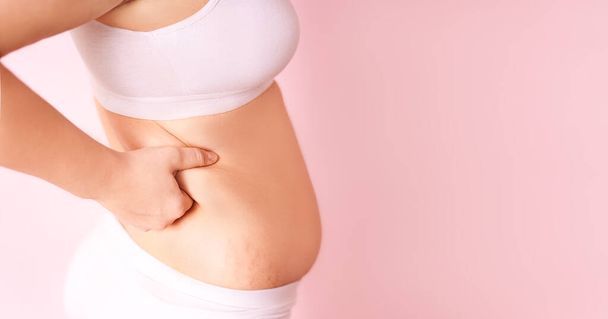 a fat girl holding a hand with excess fat on her stomach on a pink background. the concept of a womans lifestyle with a diet to reduce the belly. recovery after childbirth. place for text - Foto, Bild