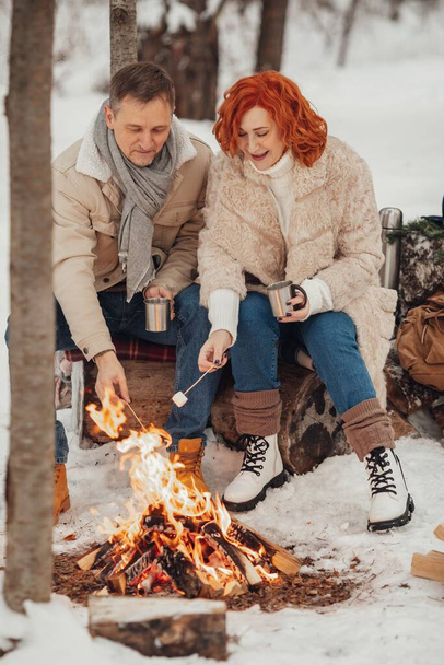 Um casal apaixonado, um homem e uma mulher em uma floresta de inverno ao redor de uma fogueira bebem café e dão marshmallows. Foto de alta qualidade - Foto, Imagem