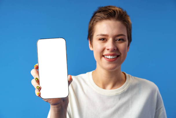 Portrait of a young woman showing blank screen mobile phone while standing over blue background - Foto, Imagen