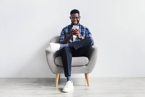 Schöner junger Schwarzer mit Handy, Surfen im Web oder in sozialen Medien, im Sessel sitzend, neue Handy-App checkend - Foto, Bild
