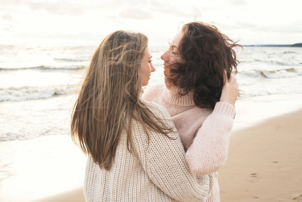 Cozy autumn walk of two girls on the shore of the windy sea. Adult sisters are trarking in blankets from the wind. A couple in love walks down the street and hugs - Photo, image