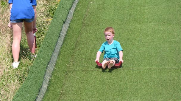 Punapää poika sininen t-paita hauskaa hauskaa nurmikolla dia Playpark aurinkoisena päivänä - Valokuva, kuva