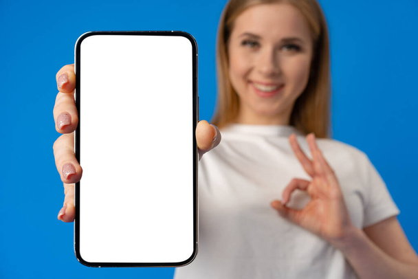 Młoda kobieta wskazuje palcem na pojedynczy ekran smartfona na tle niebieskiego koloru - Zdjęcie, obraz