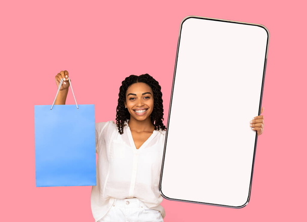 Shopping-App-Konzept. Afroamerikanerin zeigt großes Smartphone mit leerem Bildschirm und Einkaufstasche, Attrappe - Foto, Bild