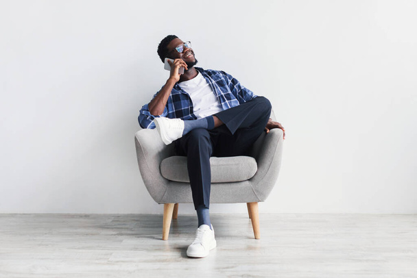 Joyeux jeune homme noir parlant sur son portable, assis dans un fauteuil, ayant une conversation téléphonique avec un ami - Photo, image