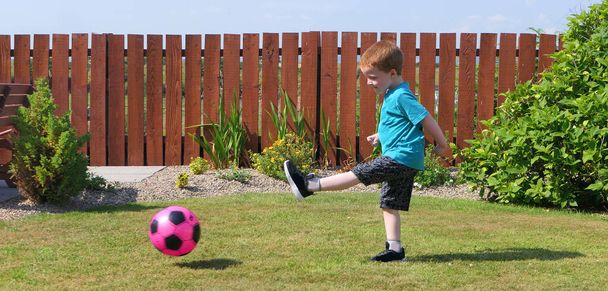 Red headed boy with a blue t-shirt having fun playing with a pink football on a lawn in a garden on a sunny day - Photo, Image