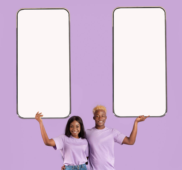 Αγαπώντας μαύρο ζευγάρι δείχνει σύγχρονα smartphones με λευκές οθόνες και χαμογελώντας πάνω από βιολετί φόντο στούντιο, mockup - Φωτογραφία, εικόνα