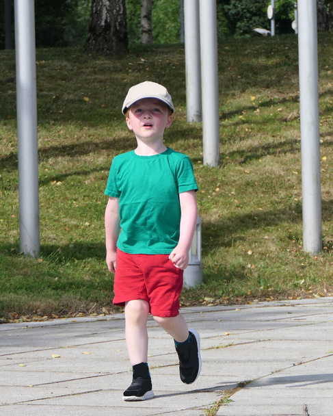 Маленький мальчик, гуляющий в парке в шляпе, зеленой футболке и красных шортах  - Фото, изображение