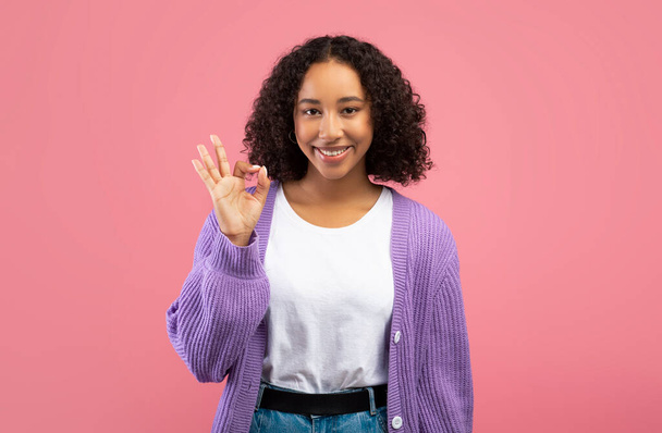 Uśmiechnięta młoda Afroamerykanka wykazująca się dobrym gestem, zatwierdzająca lub polecająca coś na różowym tle - Zdjęcie, obraz