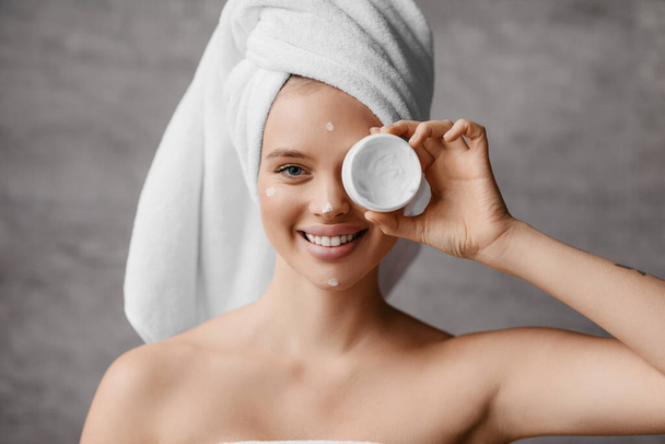 Feuchtigkeitsspendende Haut nach dem Duschen. Hübsche Millennial Frau in Handtuch bedeckt ihr Auge mit einem Glas Creme im Badezimmerinnenraum - Foto, Bild