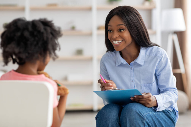 Улыбаясь, темнокожая женщина специалист по развитию ребенка разговаривает с девушкой - Фото, изображение