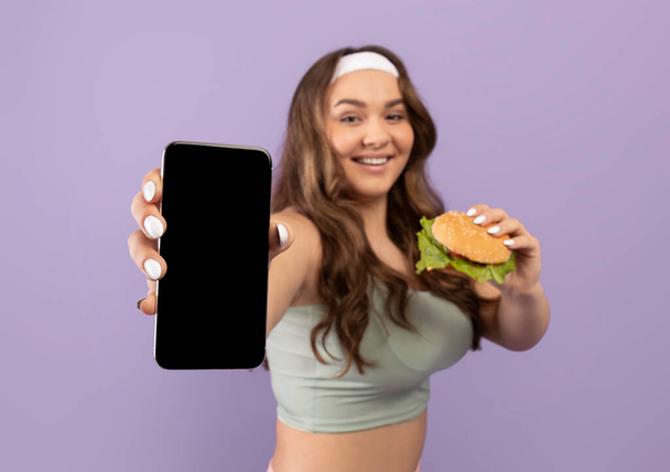 Joven alegre más mujer de tamaño en ropa deportiva con hamburguesa muestra teléfono inteligente con pantalla en blanco - Foto, Imagen