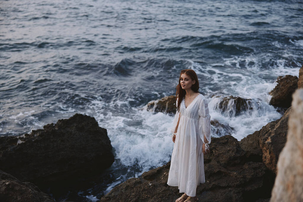 ウェディングドレスを着た女性が崖の上の海のそばに立っていて - 写真・画像