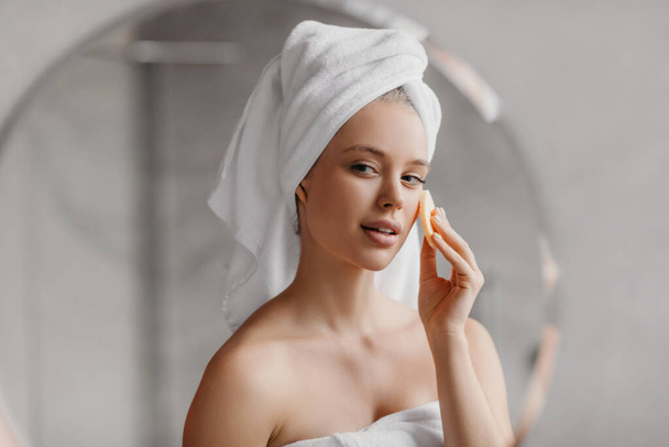 Csinos hölgy használ kozmetikai szivacs élvezi arc bőr rutin, áll a fürdőszobában reggel - Fotó, kép