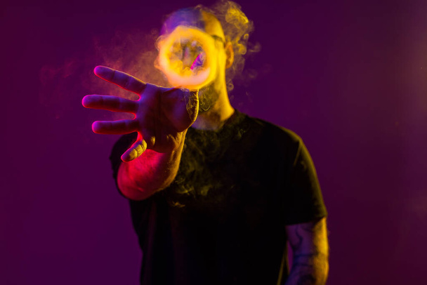 young man reaches for a ring of smoke with his hand. color filters. Vaping concept  - Фото, изображение